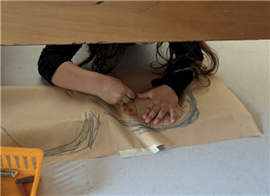 This image depicts a kindergarten student working on a cave drawing under a table.